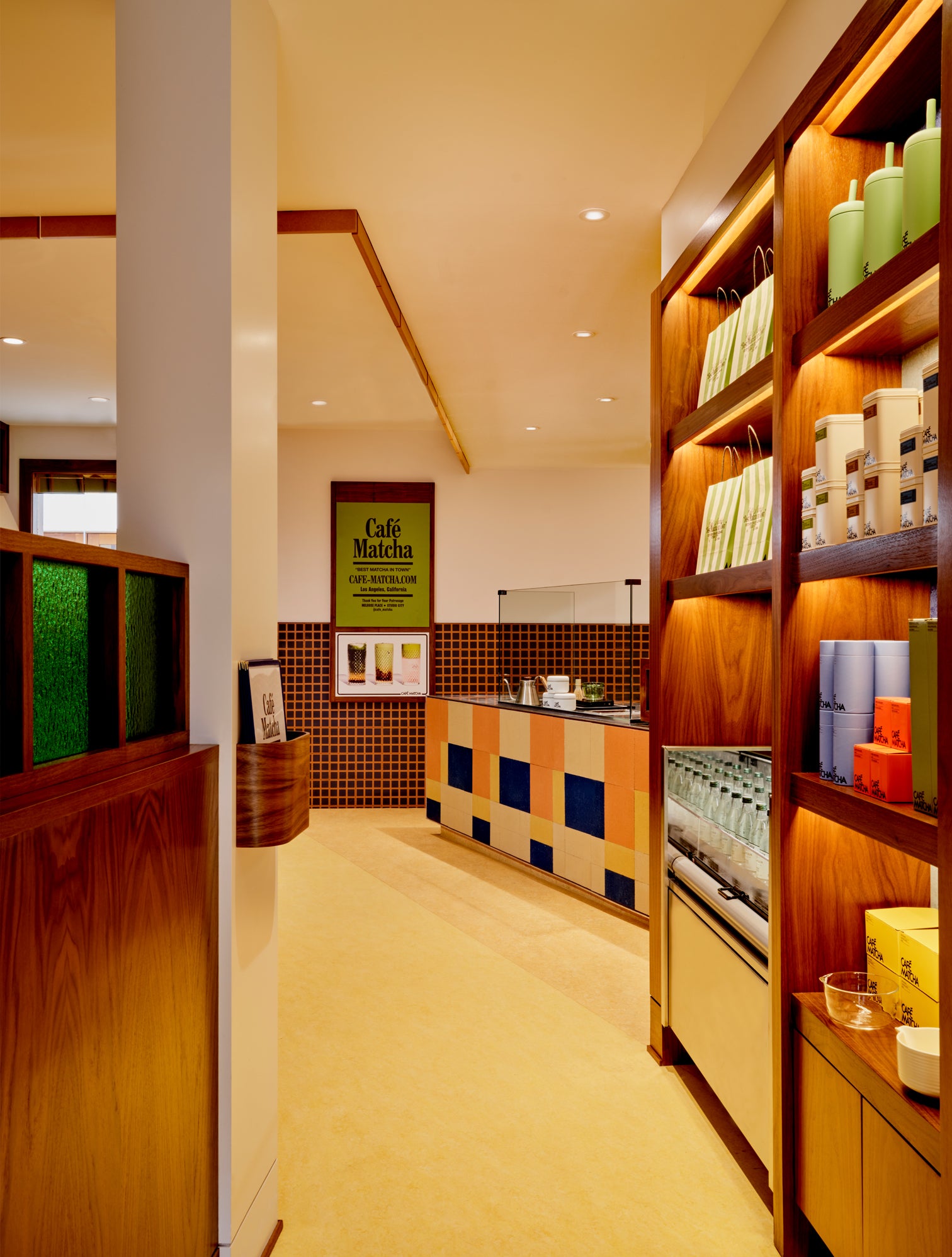 A warm, modern interior space with wooden shelving on the right displaying packaged goods, a tiled counter, and a green sign reading 'Café Matcha' in the background.
