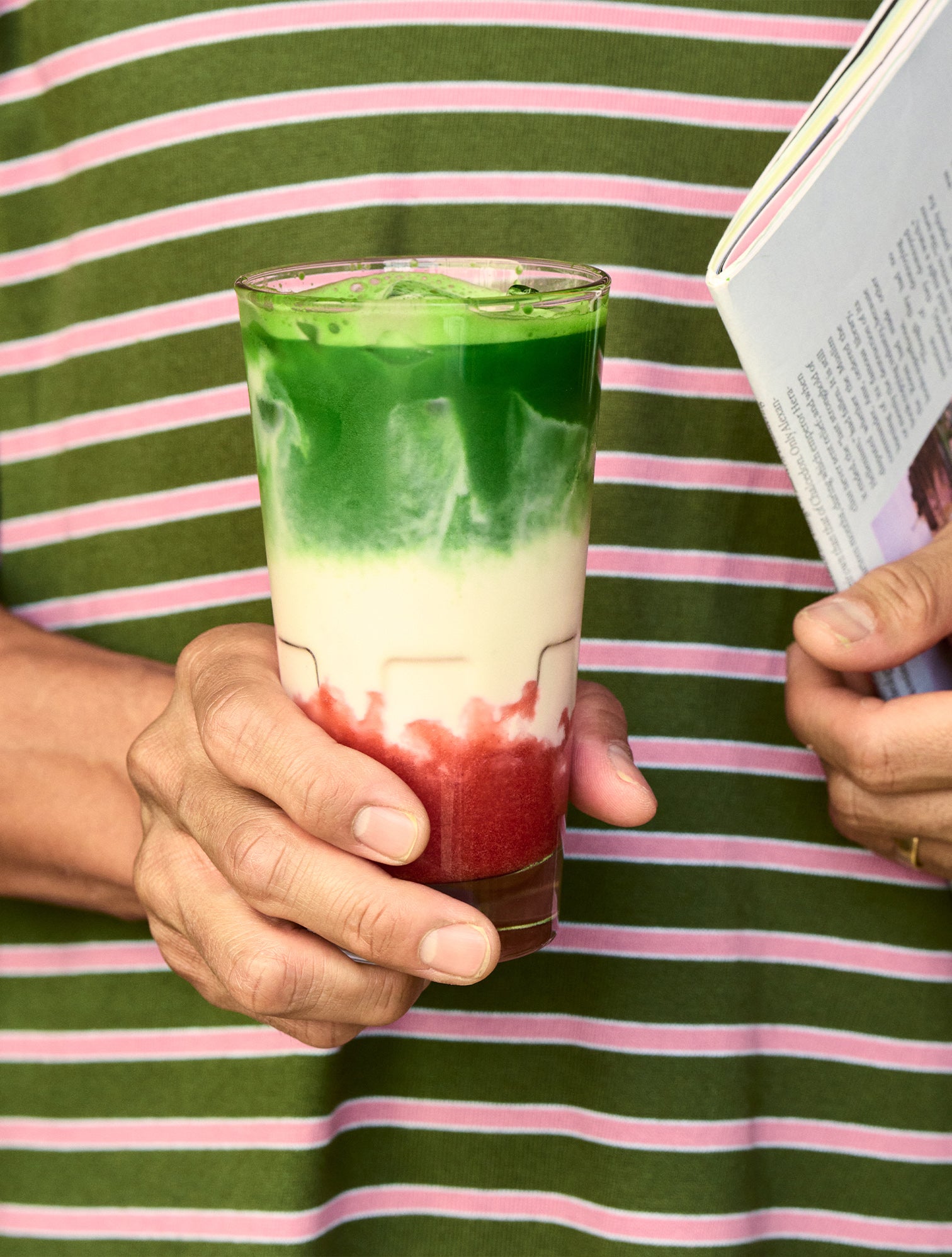 Person holding a glass with layered green, white, and red drink on a green and pink striped background